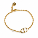Gold-tone fine chain bracelet with a polished oval letter motif and a single pearl-style bead. White background.